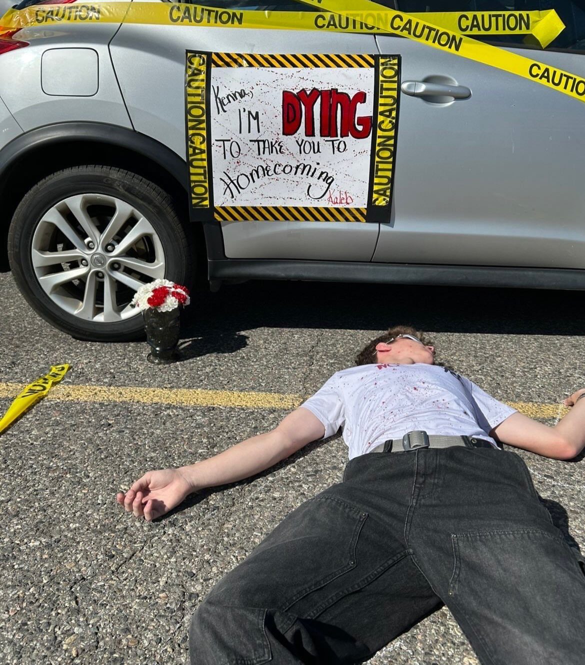 Person lying on pavement next to a car with a homecoming asking poster