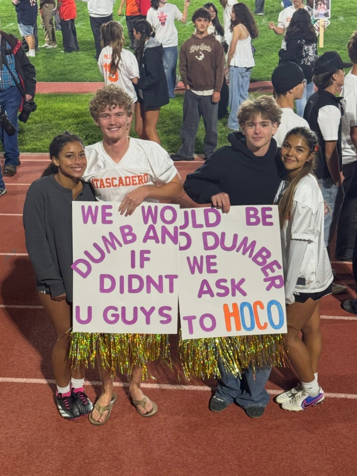 Four teenagers holding homecoming asking posters on a red athletic running track.
