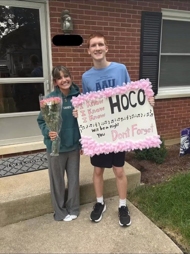 Teenage couple posing outside with a music-themed homecoming poster and pink roses.