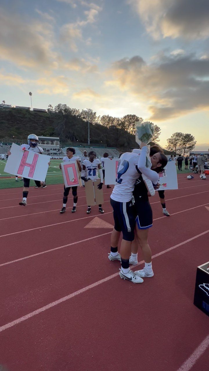 Two athletes hugging on a track while teammates hold HOCO asking posters behind them.
