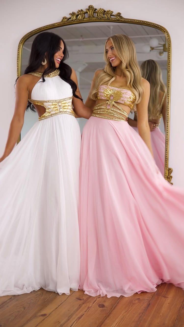 Two women in white and pink formal gowns with gold accents posing by a mirror.