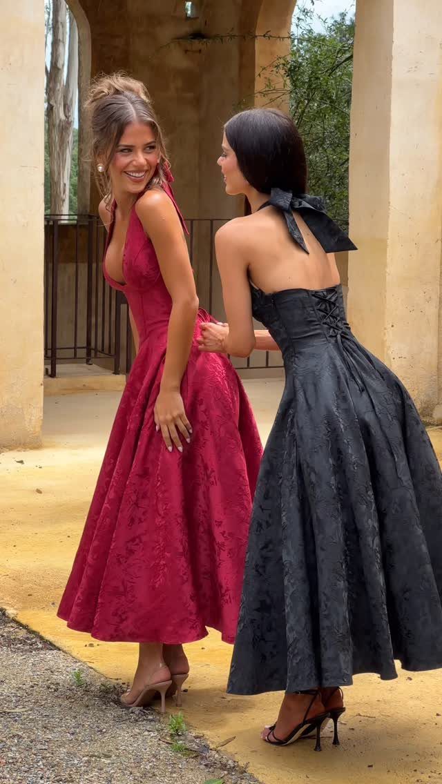 Two young women wearing tea-length red and black formal dresses standing outdoors.