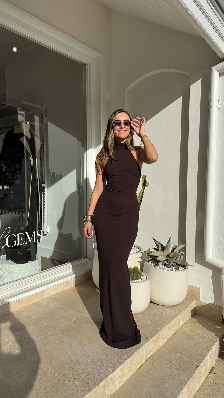 Woman in a long dark brown high-neck sleeveless dress standing outdoors by a store.