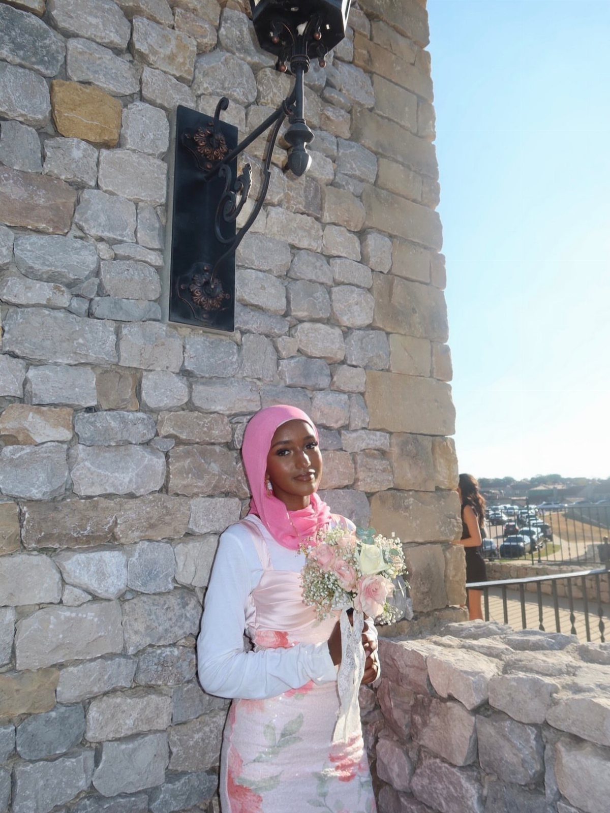 Woman in pink hijab and floral dress holding a bouquet by a stone wall