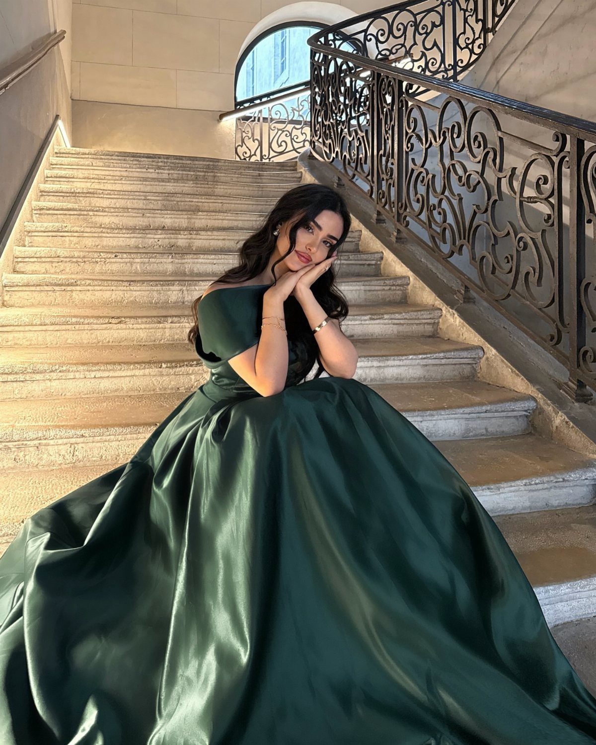 Woman in a voluminous dark green satin gown sitting on stone stairs.