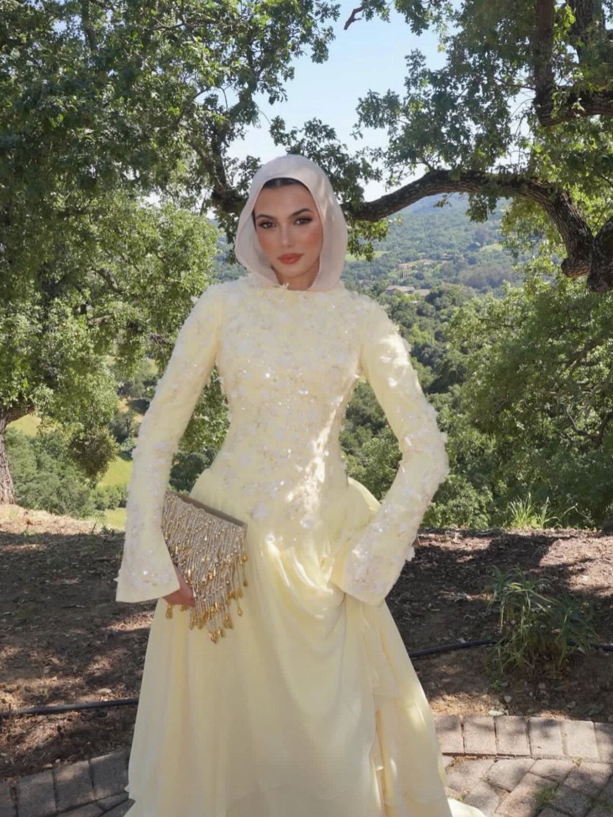 Woman in a pale yellow modest long-sleeved gown and hijab standing outdoors.