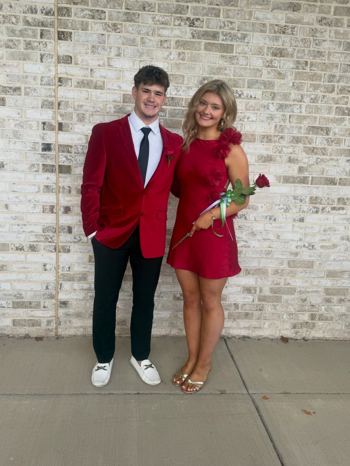 Young couple in matching red formal attire posing against a brick wall.