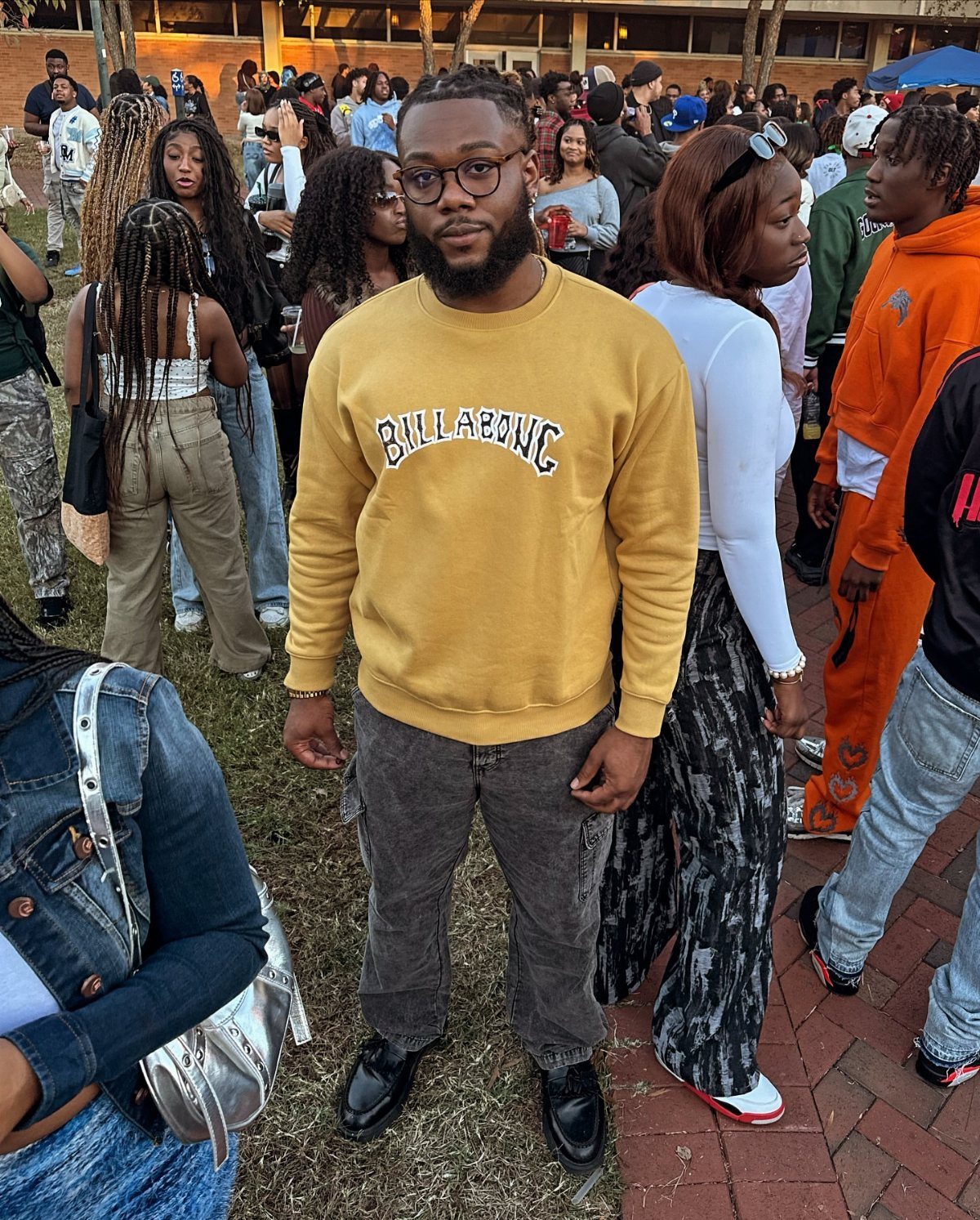 Man wearing yellow sweatshirt and grey cargo pants standing in a crowd outdoors.