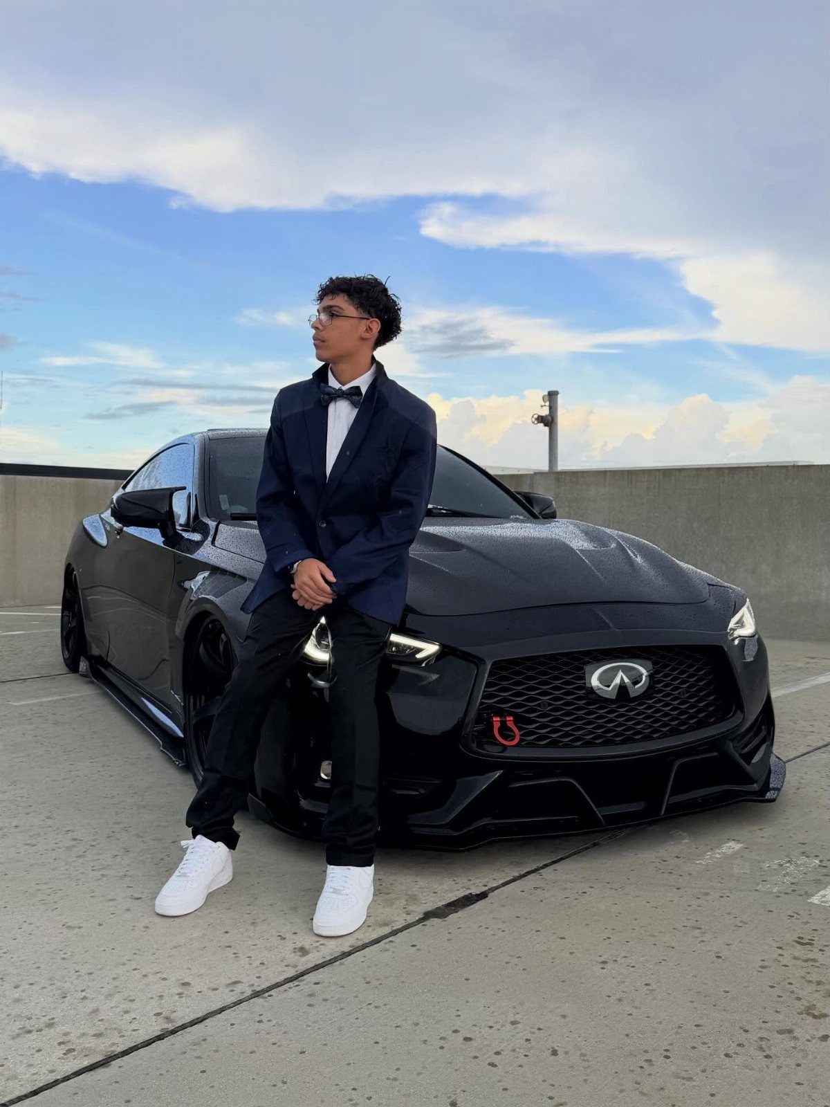 Young man in navy blazer and bow tie posing by black sports car.