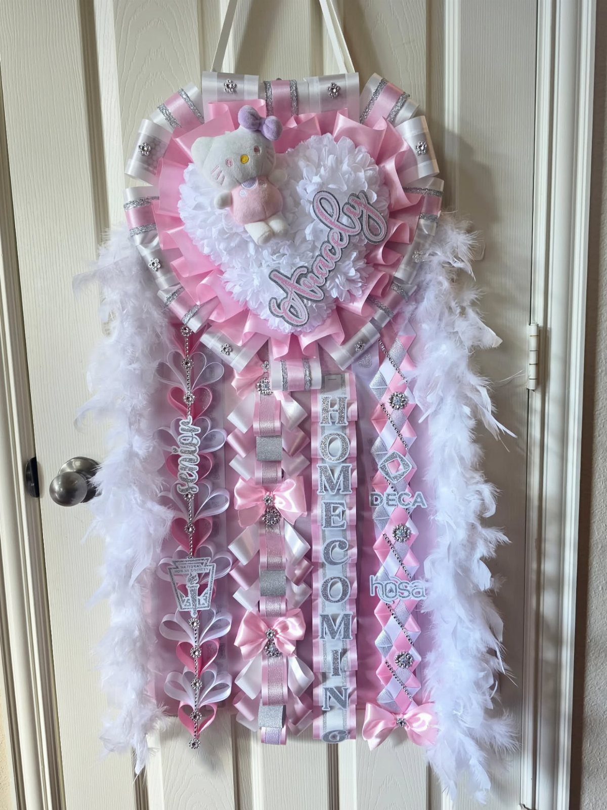 Hello Kitty senior homecoming mum with pink ribbons and white feathers on a door