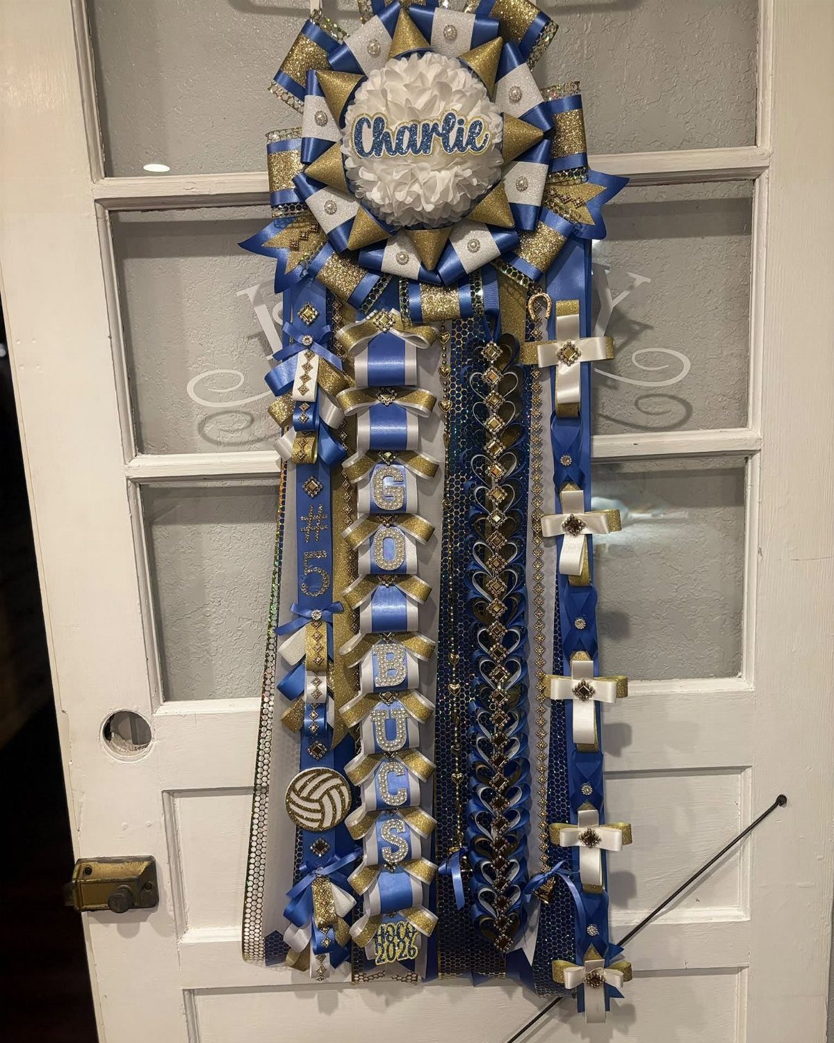Ornate blue and gold homecoming mum with name ribbons hanging on white door