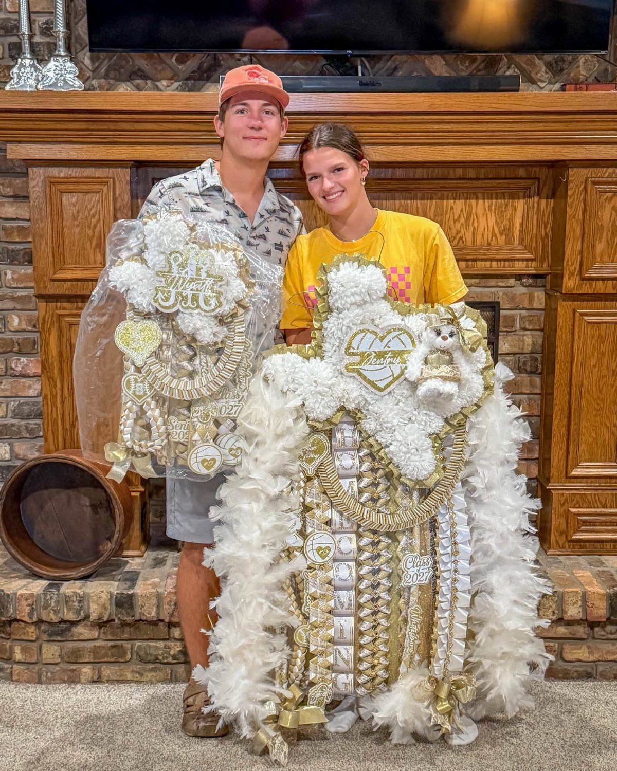 Teenage boy and girl wearing large white and gold homecoming mums by a fireplace