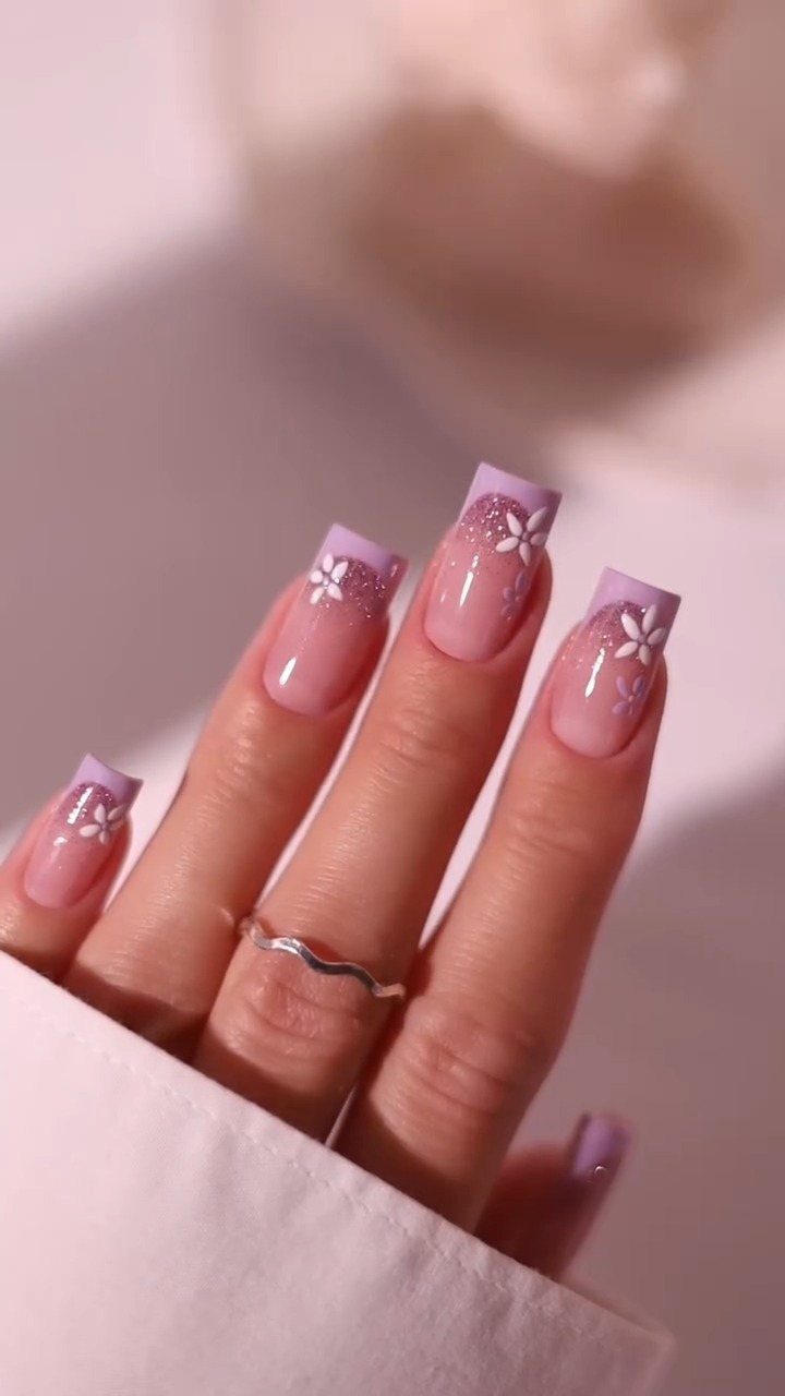 Long square nails with purple tips, white flowers, and glitter on a hand.