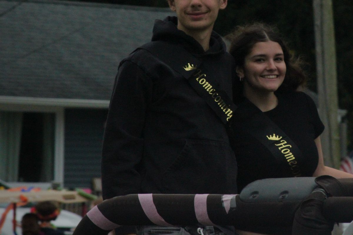 Teenage boy and girl wearing black clothes and gold homecoming sashes smiling outdoors.