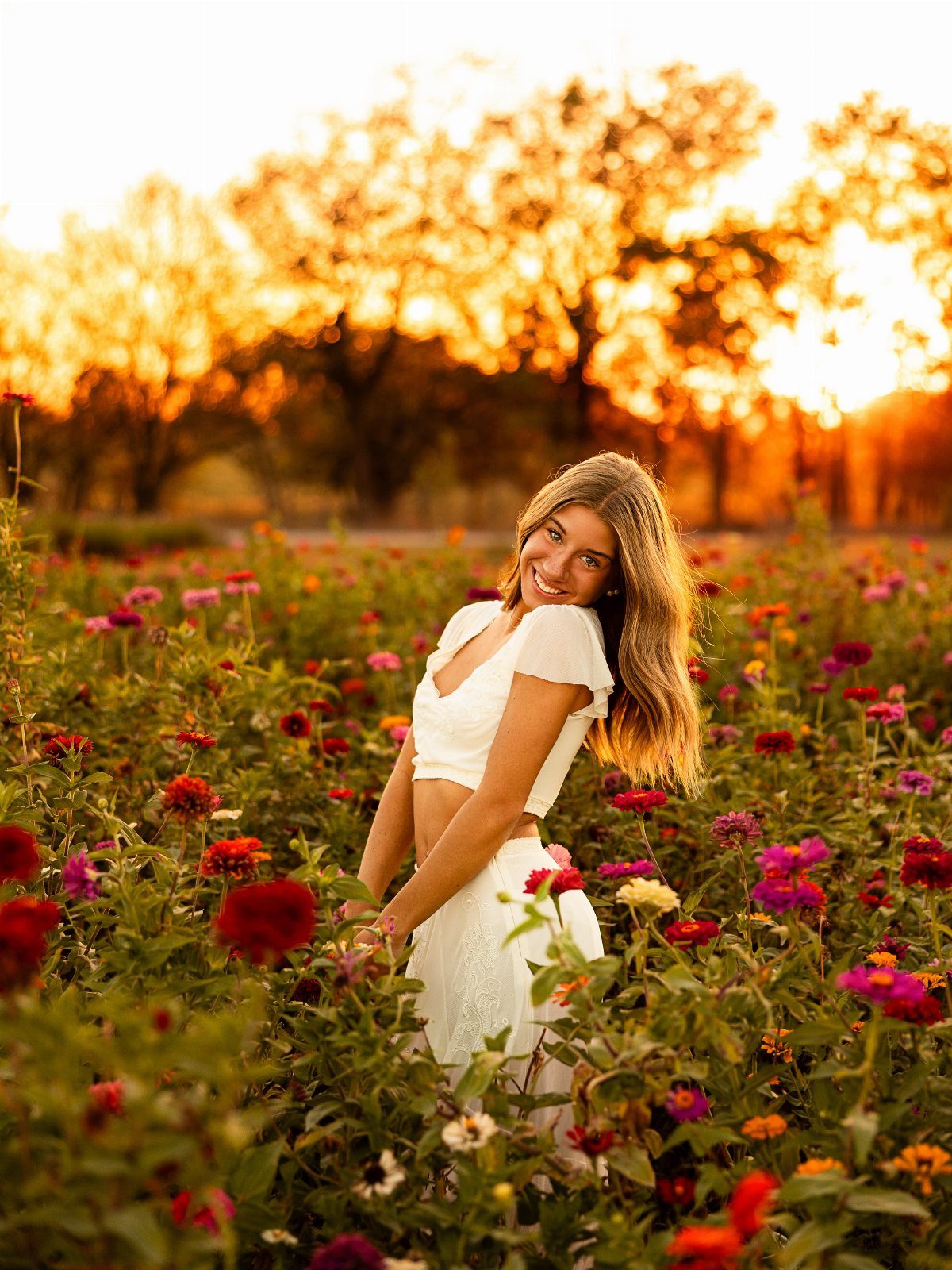Young woman in white outfit smiling in a colorful flower field at sunset
