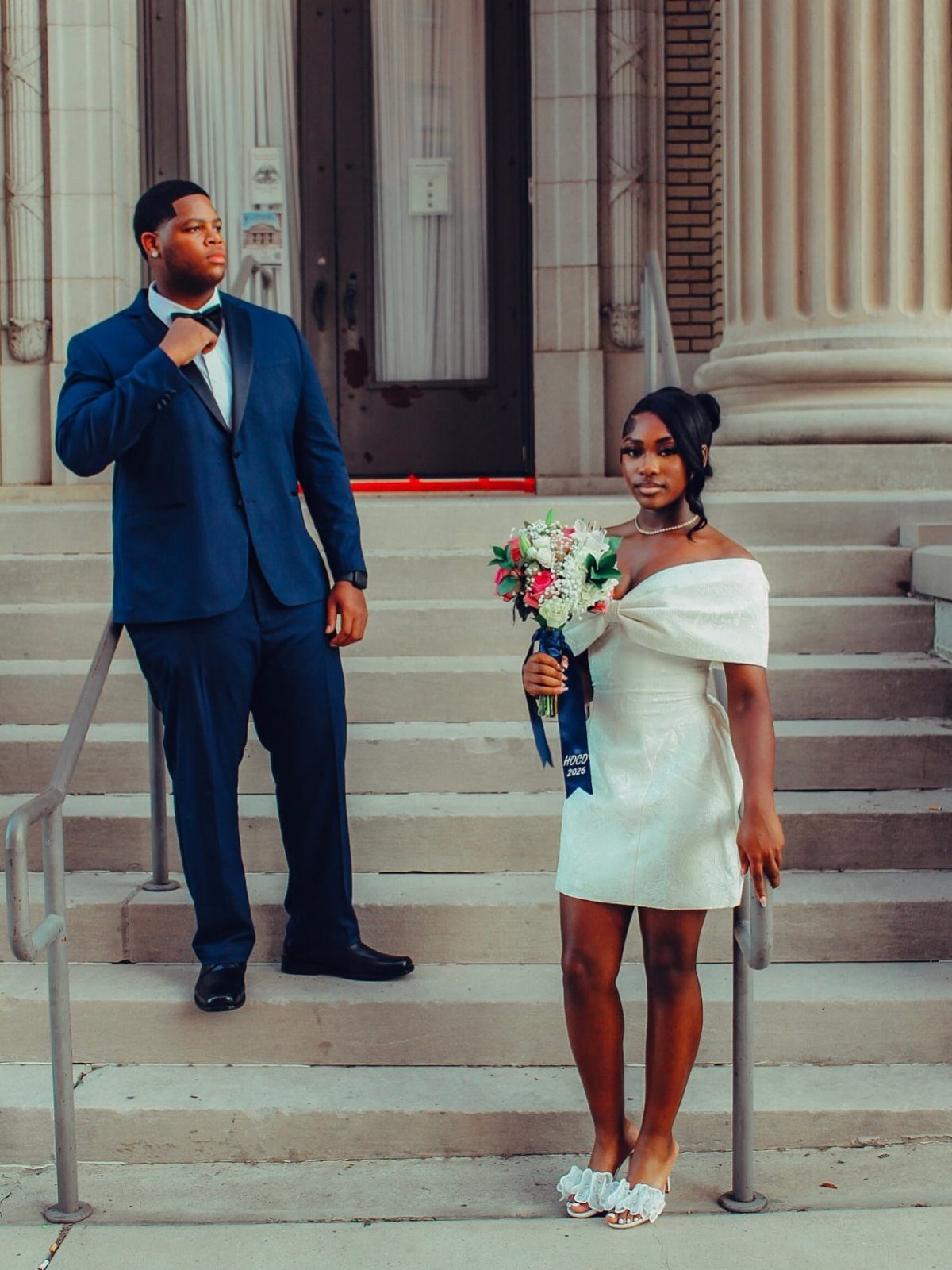 Couple in formal attire posing on concrete stairs with a floral bouquet.