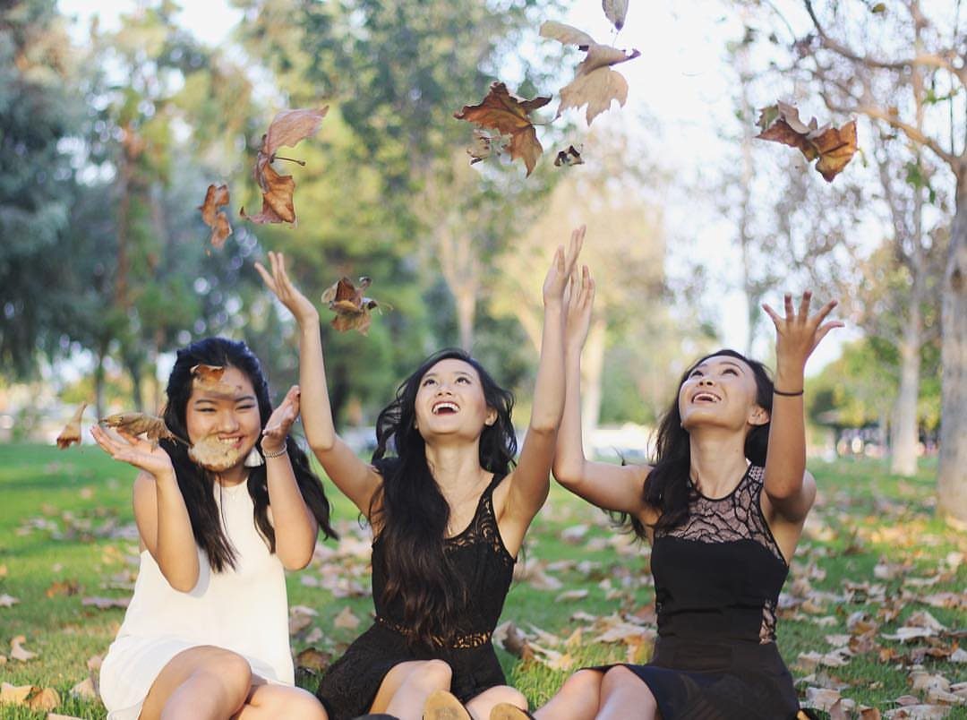 Three young women in dresses tossing autumn leaves while sitting on green grass.