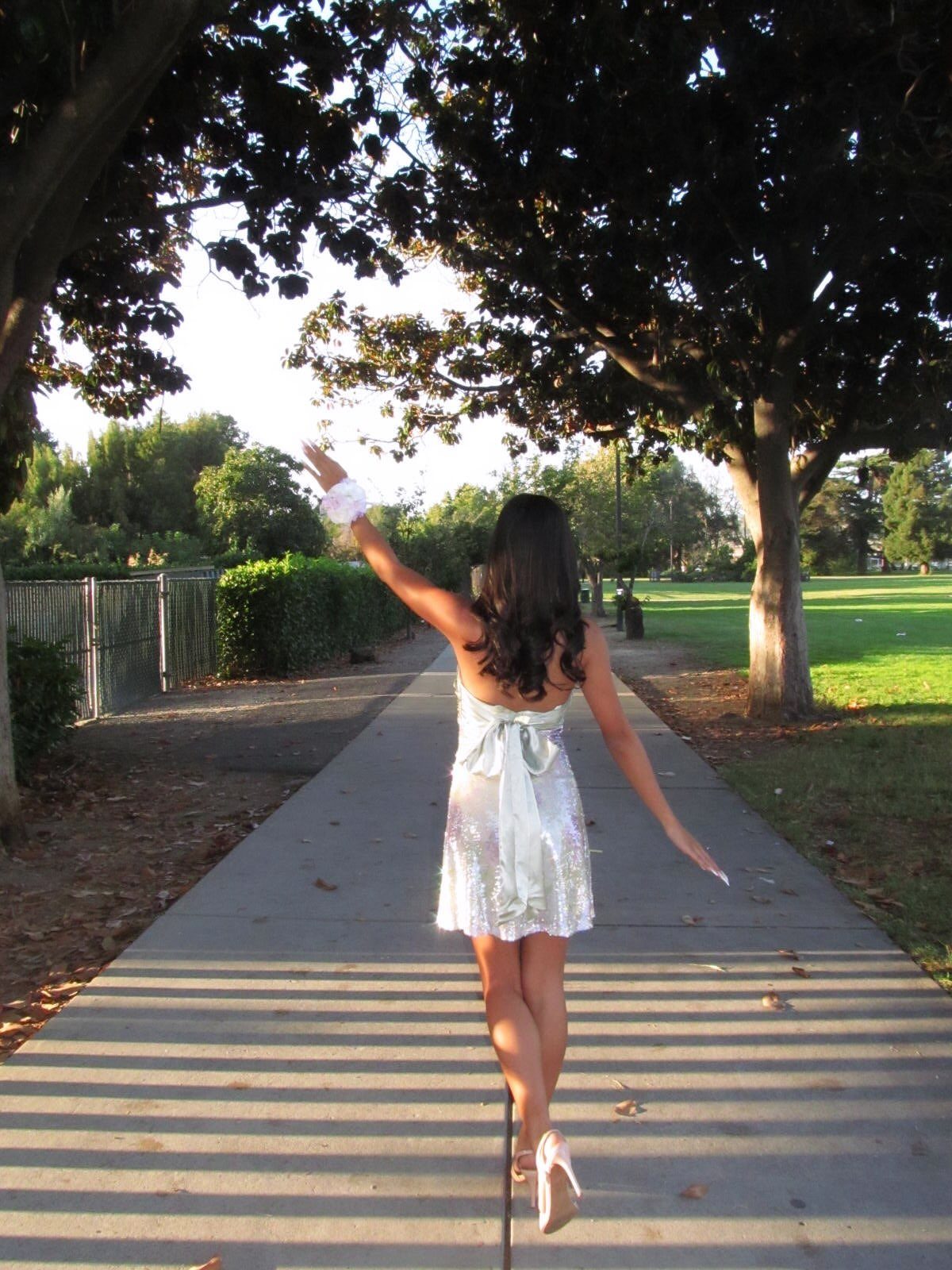 Woman in a silver sequin dress walking away on a sunny park path.