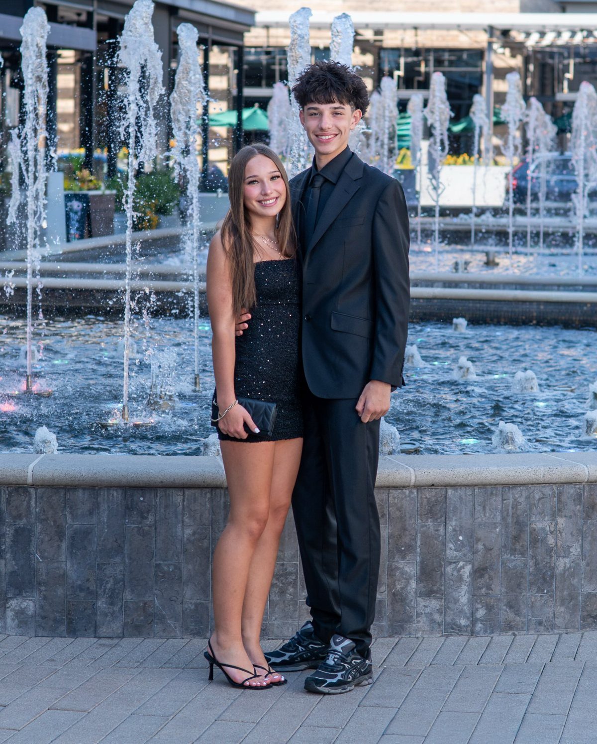 Teenage couple in formal black attire posing before a splashing outdoor water fountain.