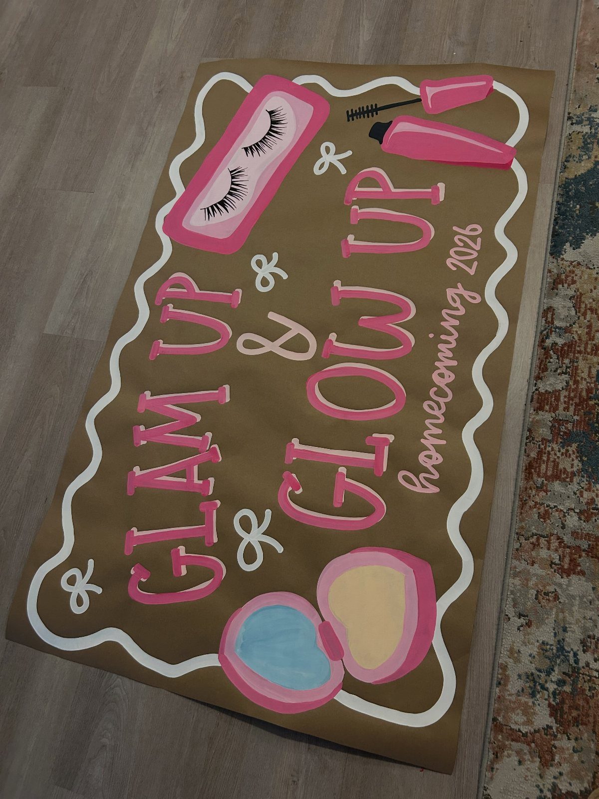 Brown homecoming poster with pink lettering and makeup illustrations on a wooden floor.