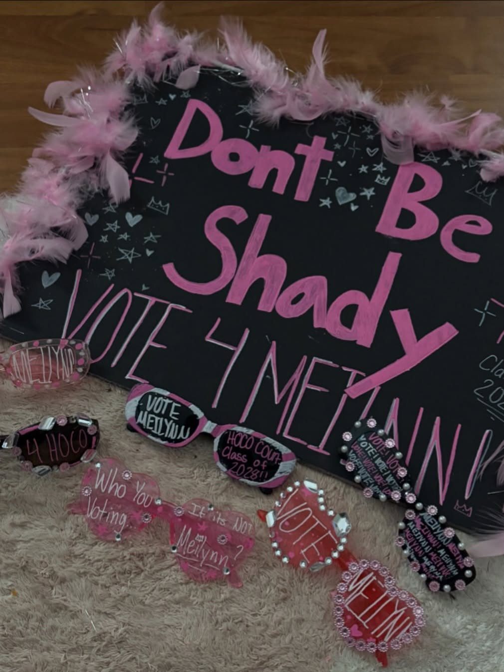 Black poster with pink text and feathers encouraging votes for Meillynn with decorated glasses.