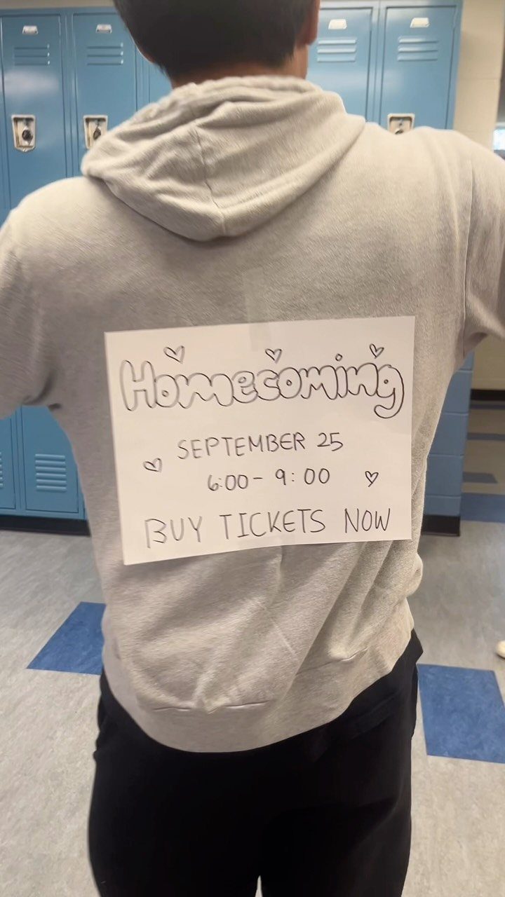 Person wearing a grey hoodie with a handmade homecoming announcement sign taped to back