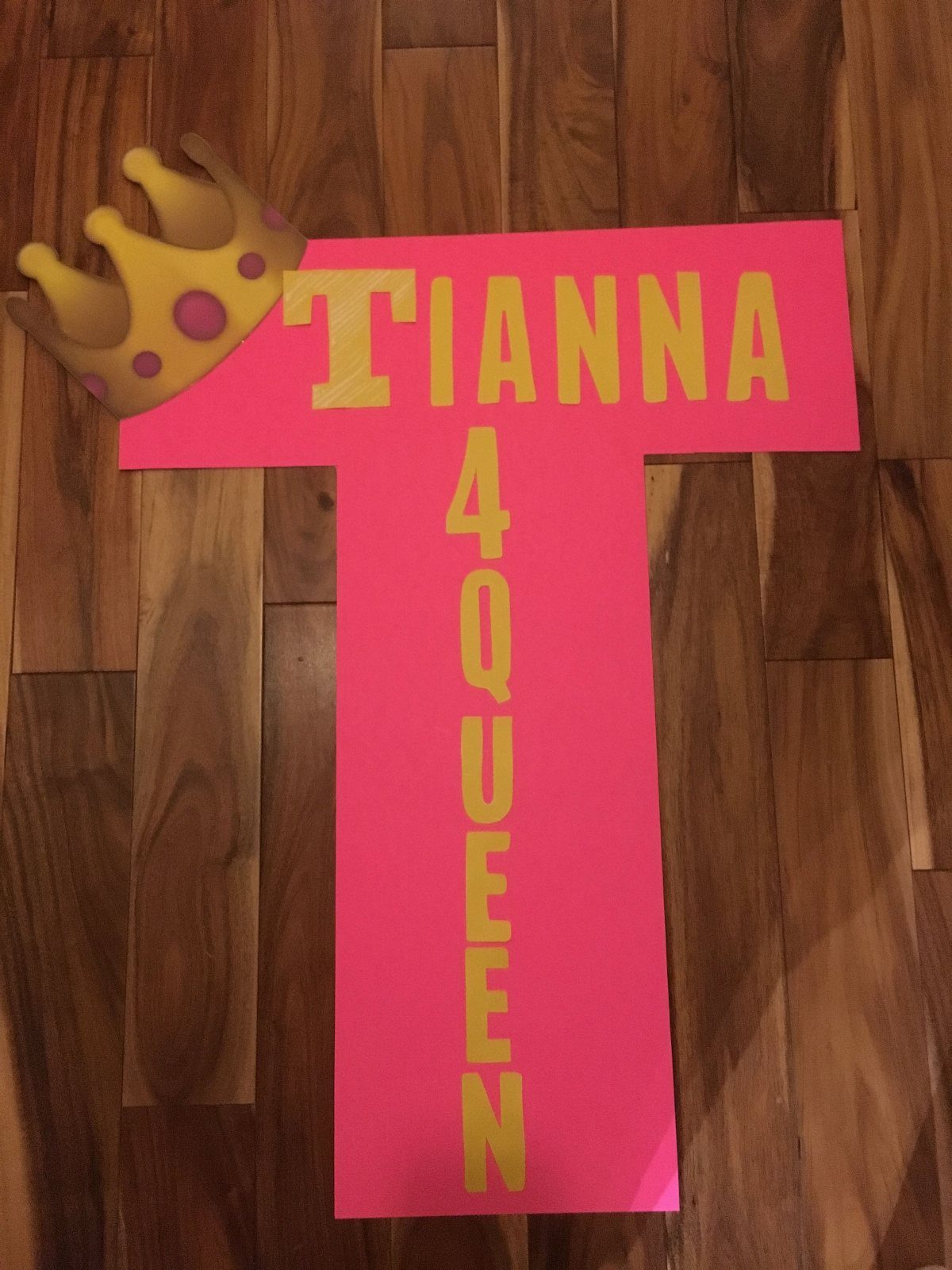 Pink T-shaped poster board with yellow text and a crown on wooden floor