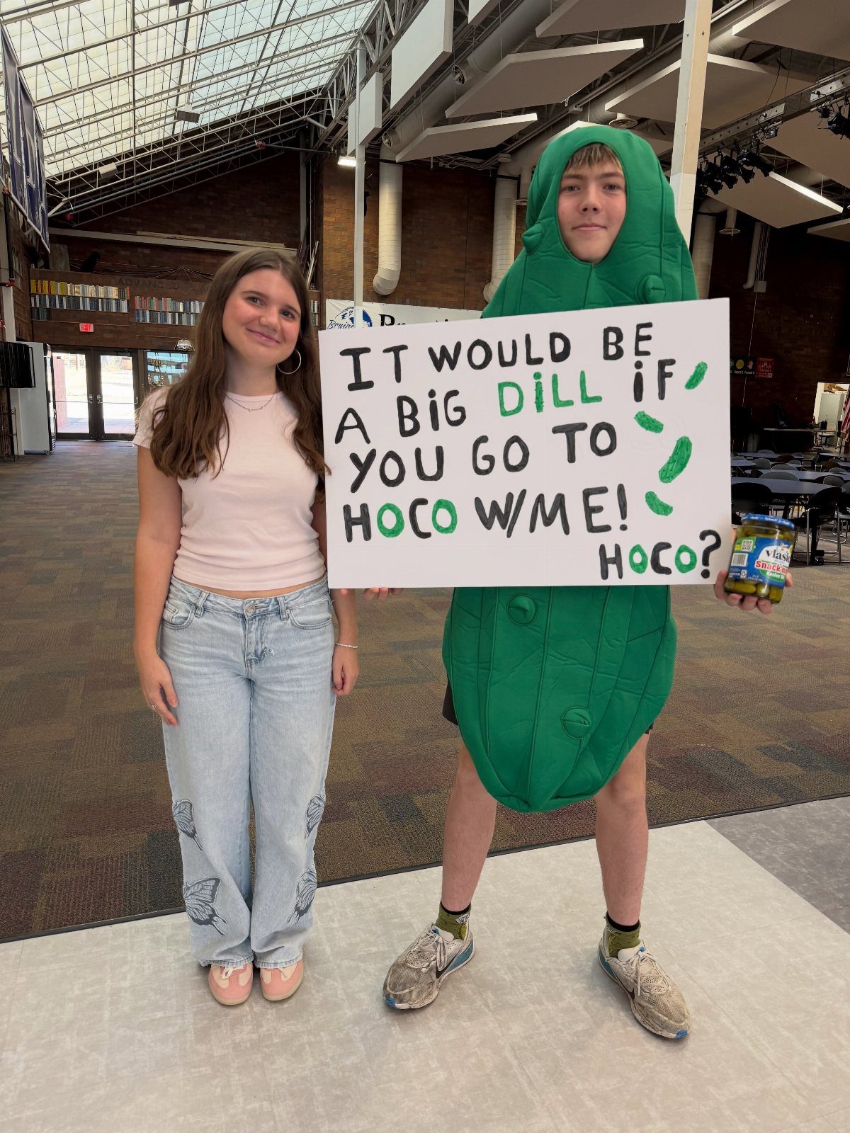 Teenage boy in a pickle costume holding a homecoming proposal sign next to a girl.