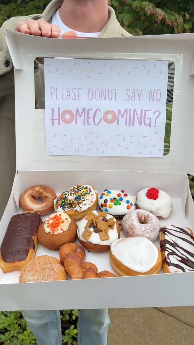 Open donut box with a homecoming proposal sign and assorted colorful donuts.
