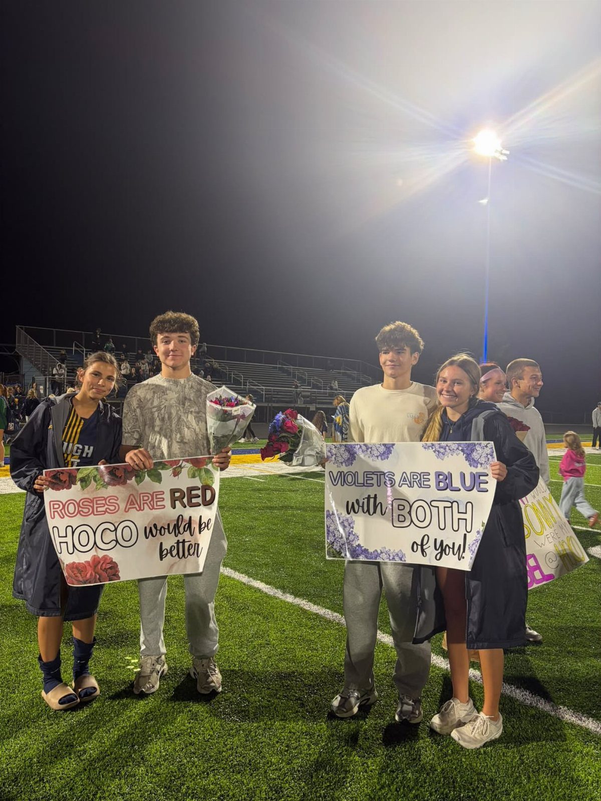 Four teenagers on a football field holding posters for a homecoming proposal.
