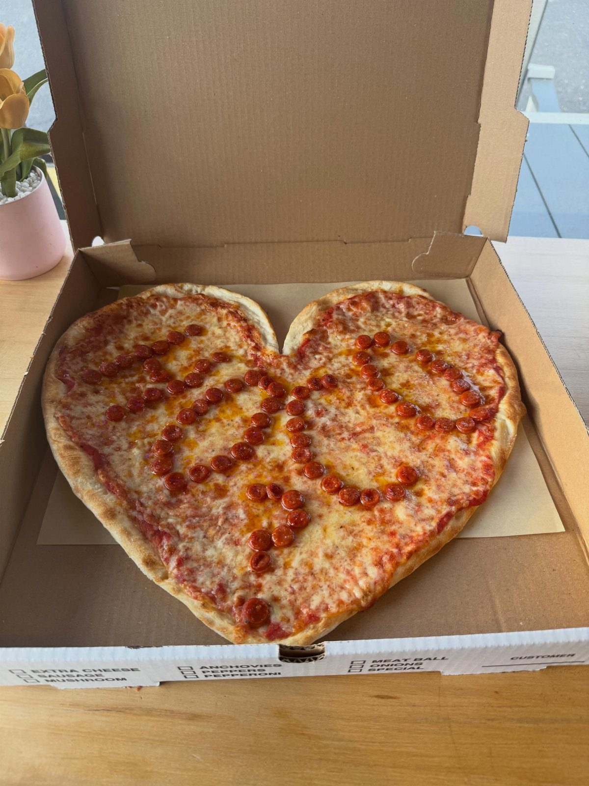 Heart-shaped pizza with pepperoni spelling out HOCO? inside a cardboard delivery box