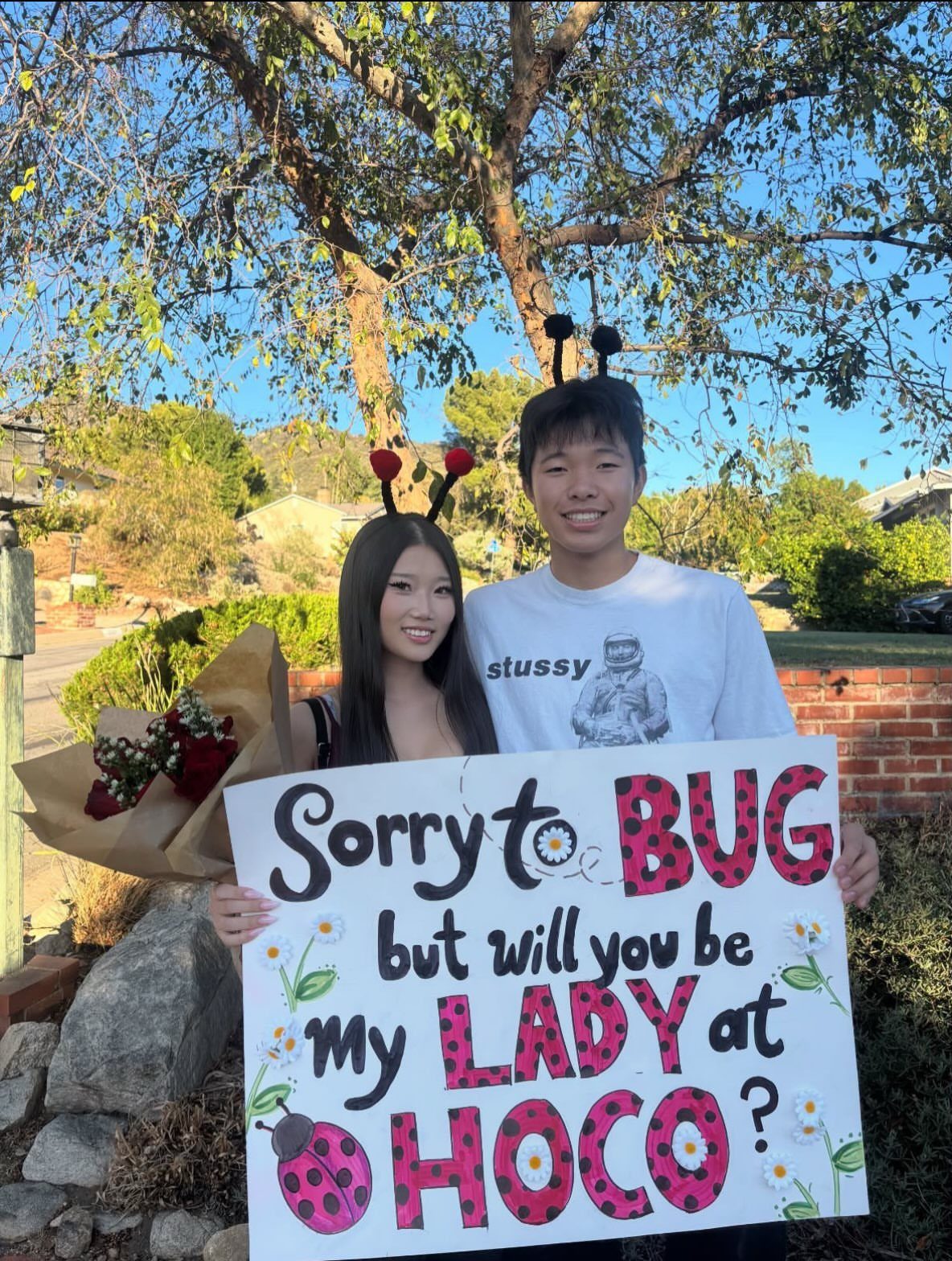 Teenage couple posing with a ladybug themed homecoming proposal poster and red roses.