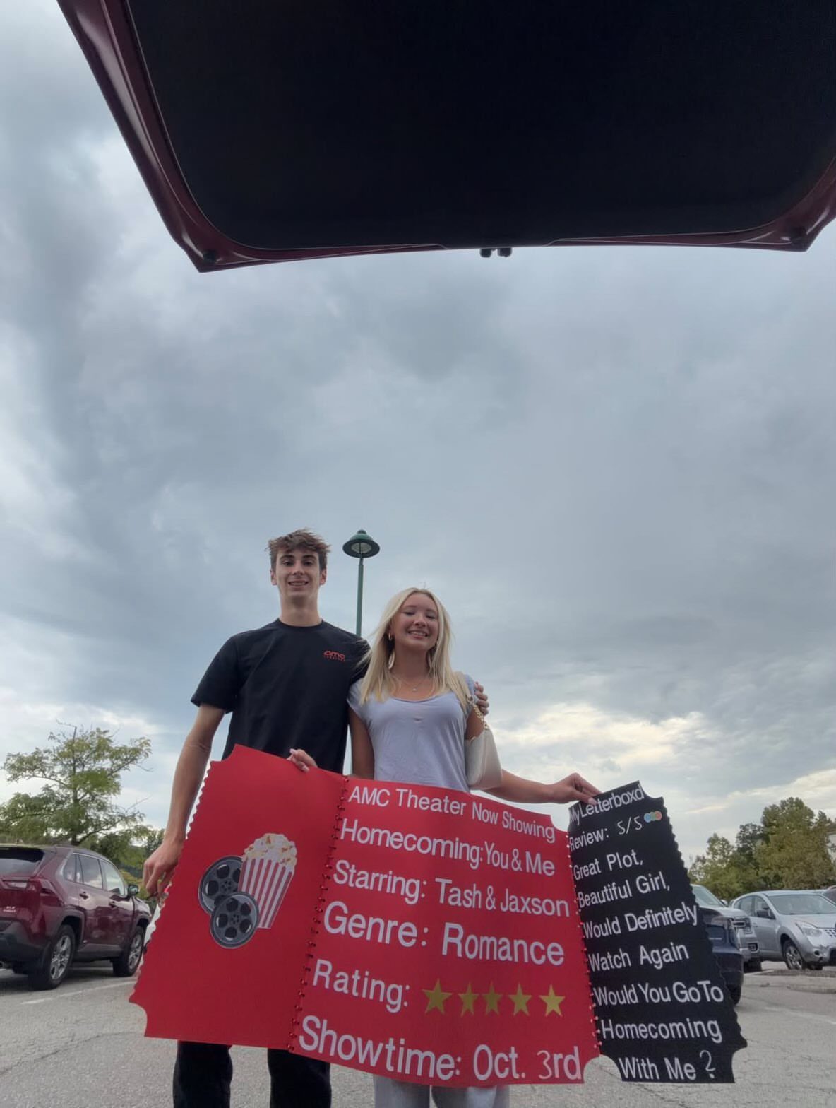 Teenage couple holding a large red movie ticket poster for a homecoming proposal.