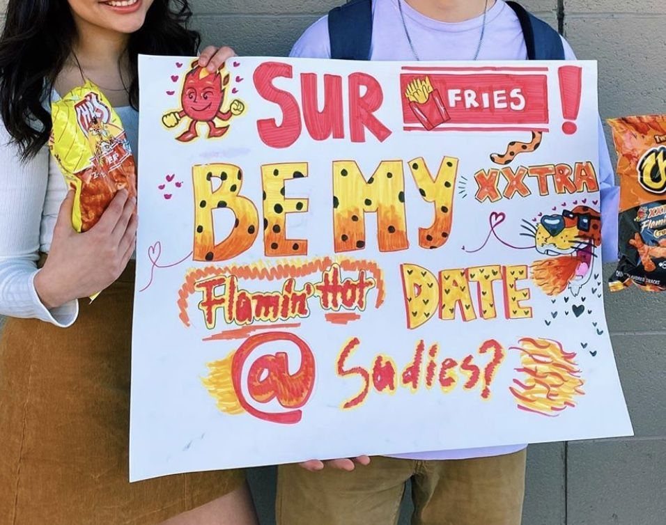 Two people holding a colorful homecoming proposal sign with spicy snack food puns.