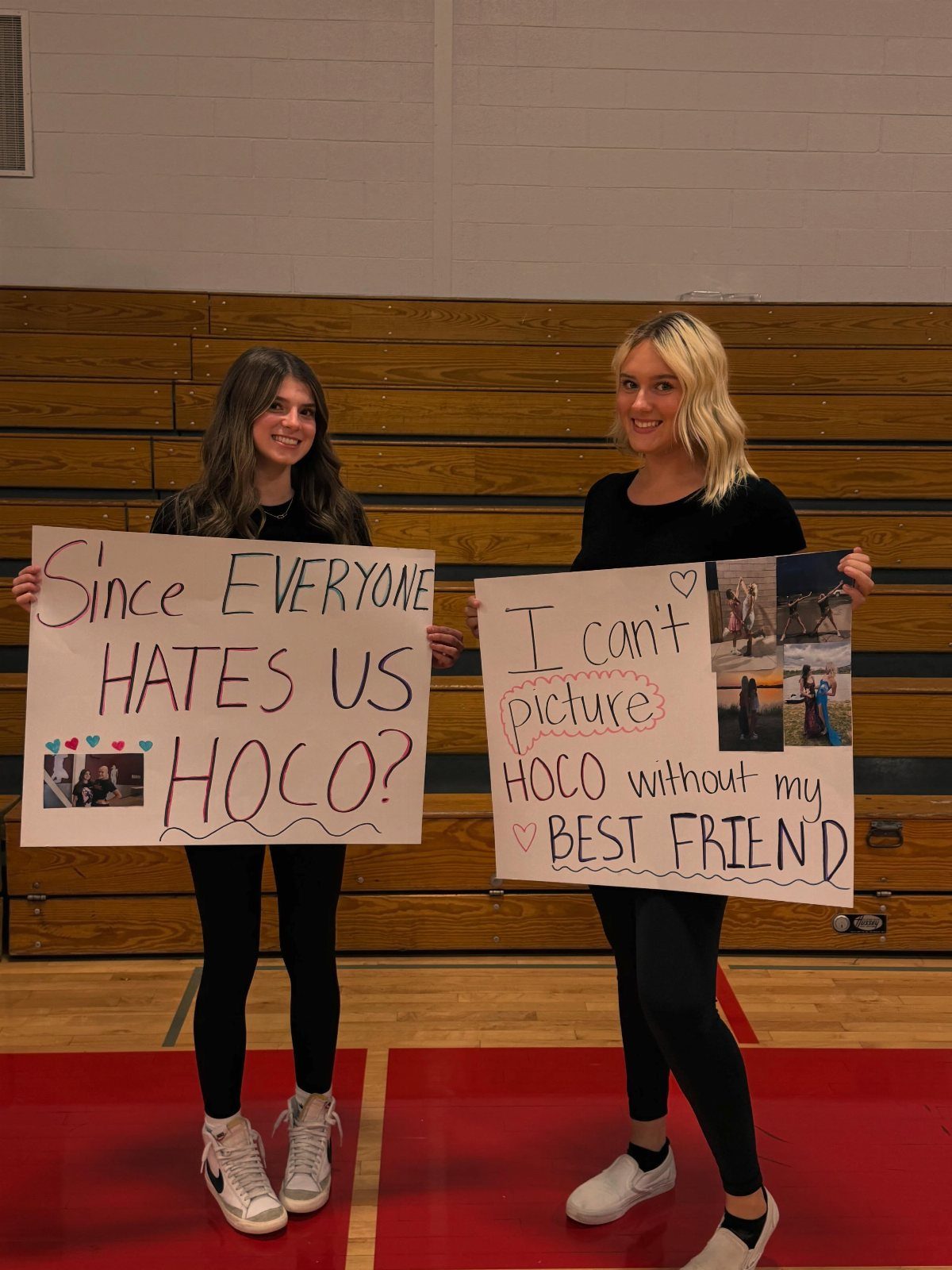 Two girls holding homecoming proposal signs in a gym with wooden bleachers.