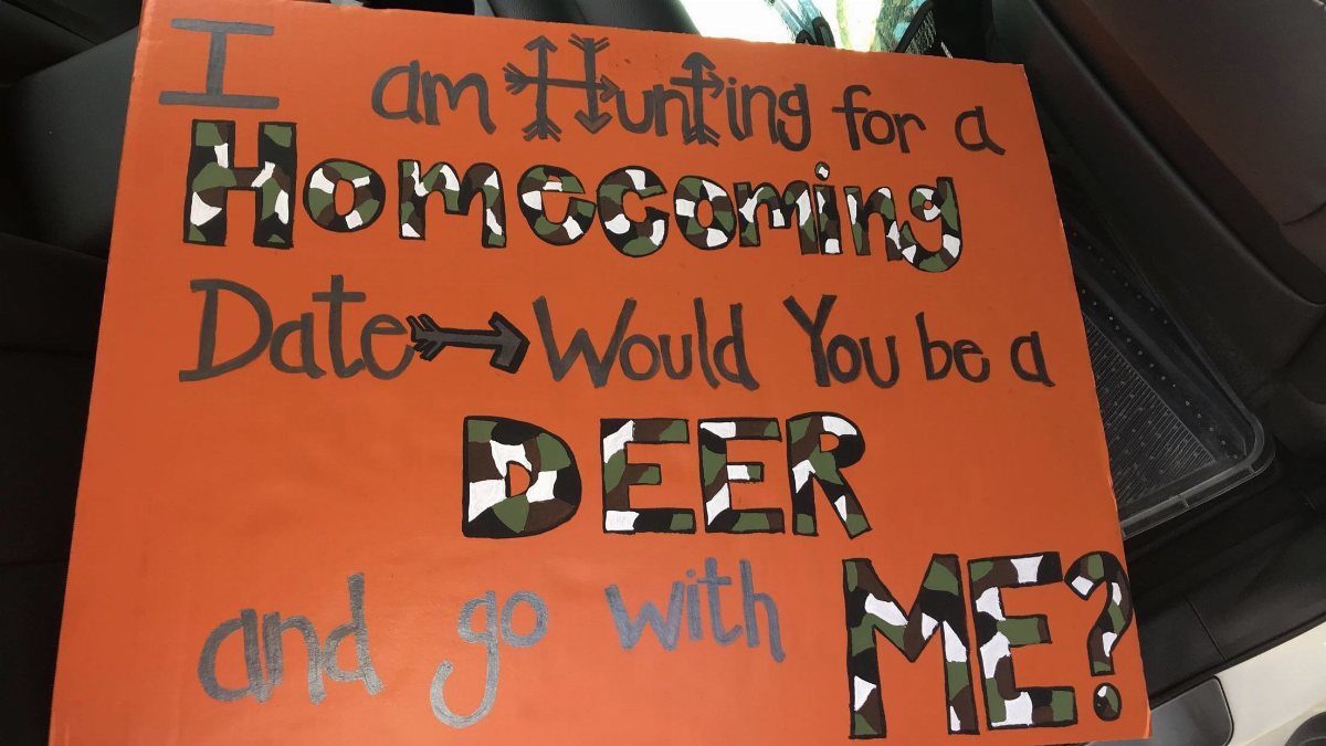Orange homecoming proposal sign with hunting puns and camouflage lettering inside a car