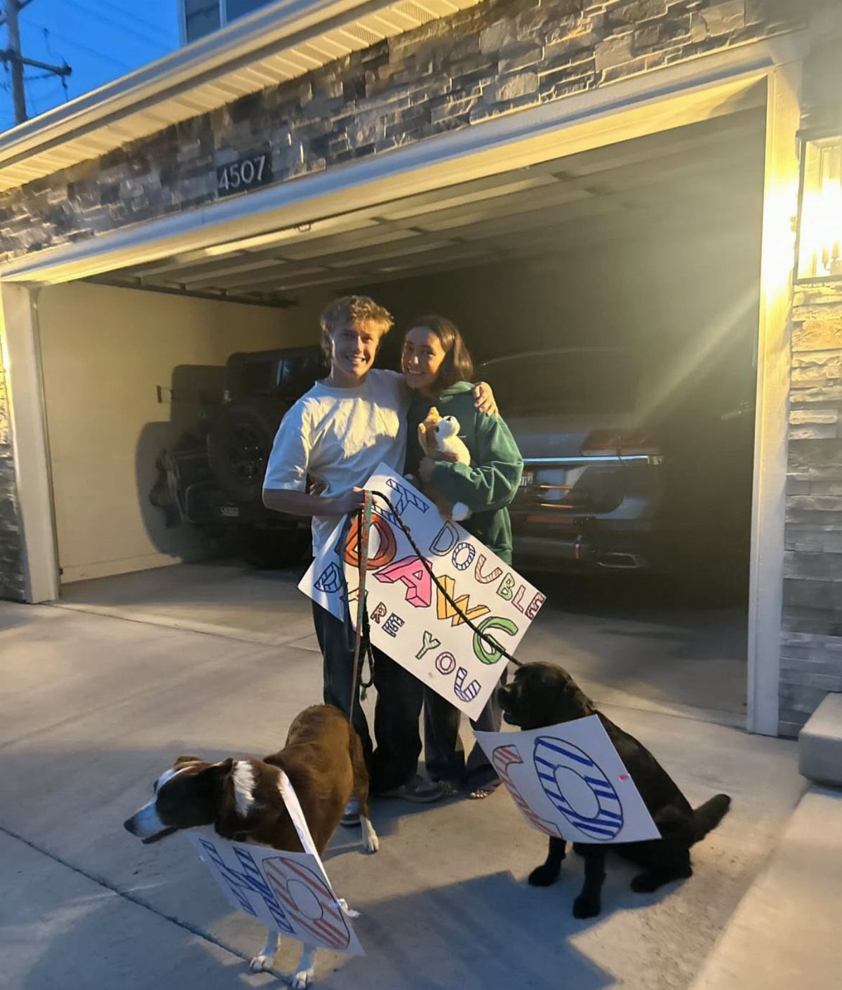 Teenage couple with two dogs wearing signs for a homecoming proposal in a driveway