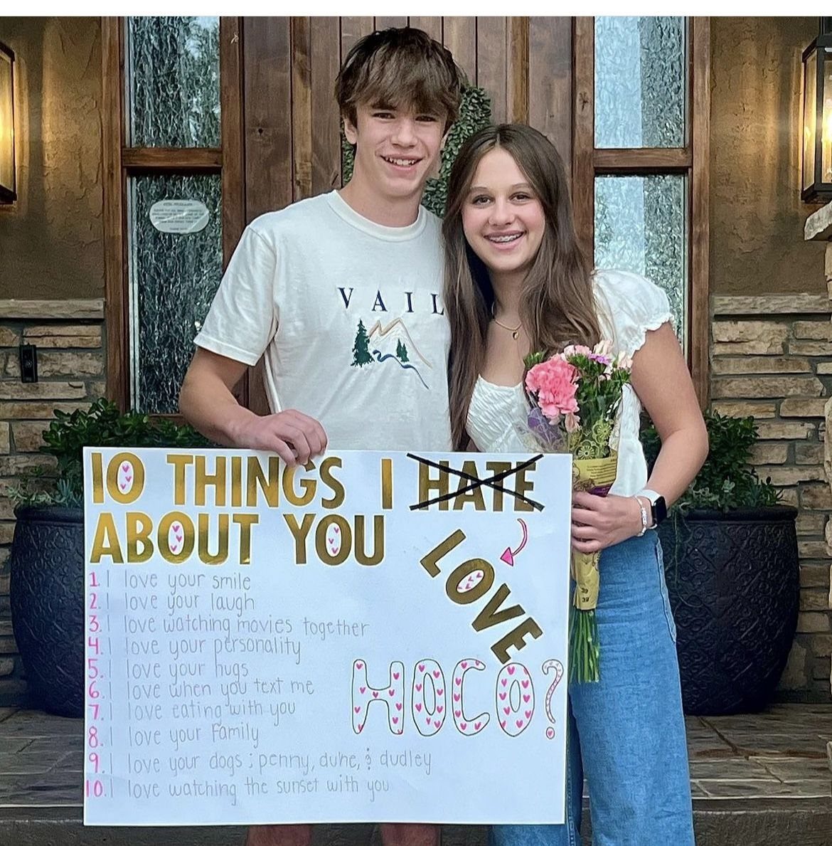 Teenage couple posing with a movie-themed homecoming proposal sign and a bouquet of flowers.