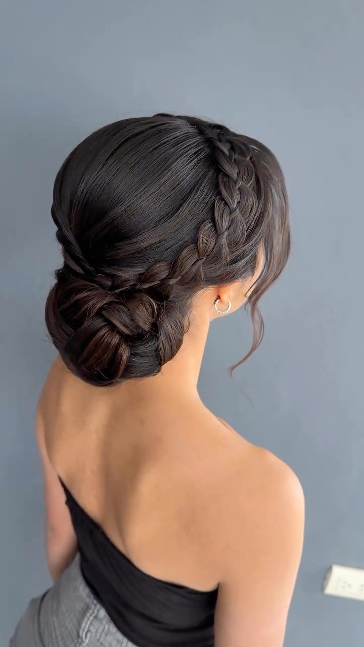 Woman with dark hair in a braided updo and low bun against grey wall
