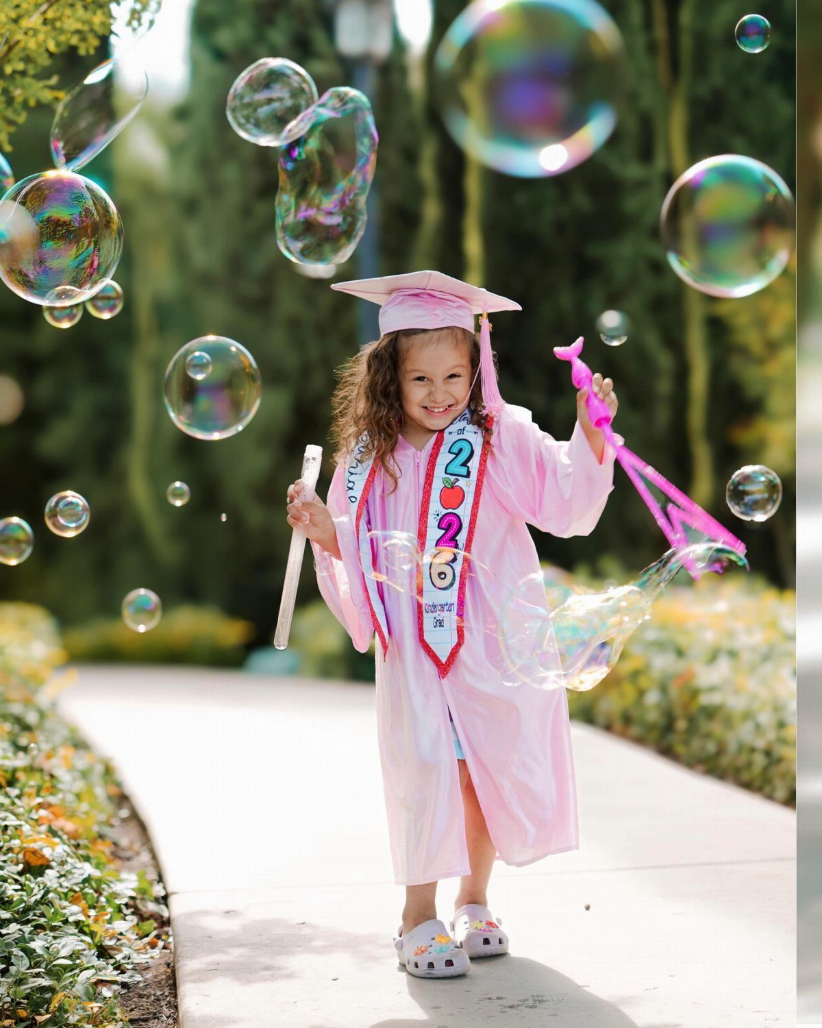 Young girl in pink graduation gown and cap smiling among floating bubbles.