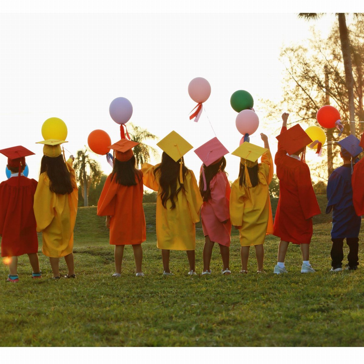 Children in colorful graduation gowns and caps holding balloons in a grassy field