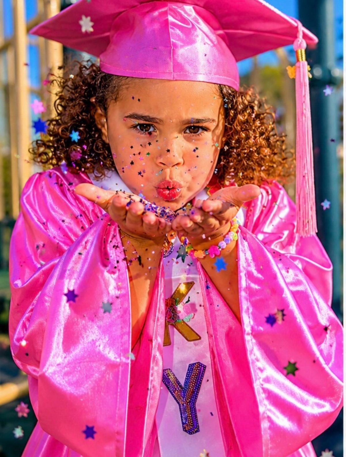 Young girl in pink graduation cap and gown blowing colorful star confetti