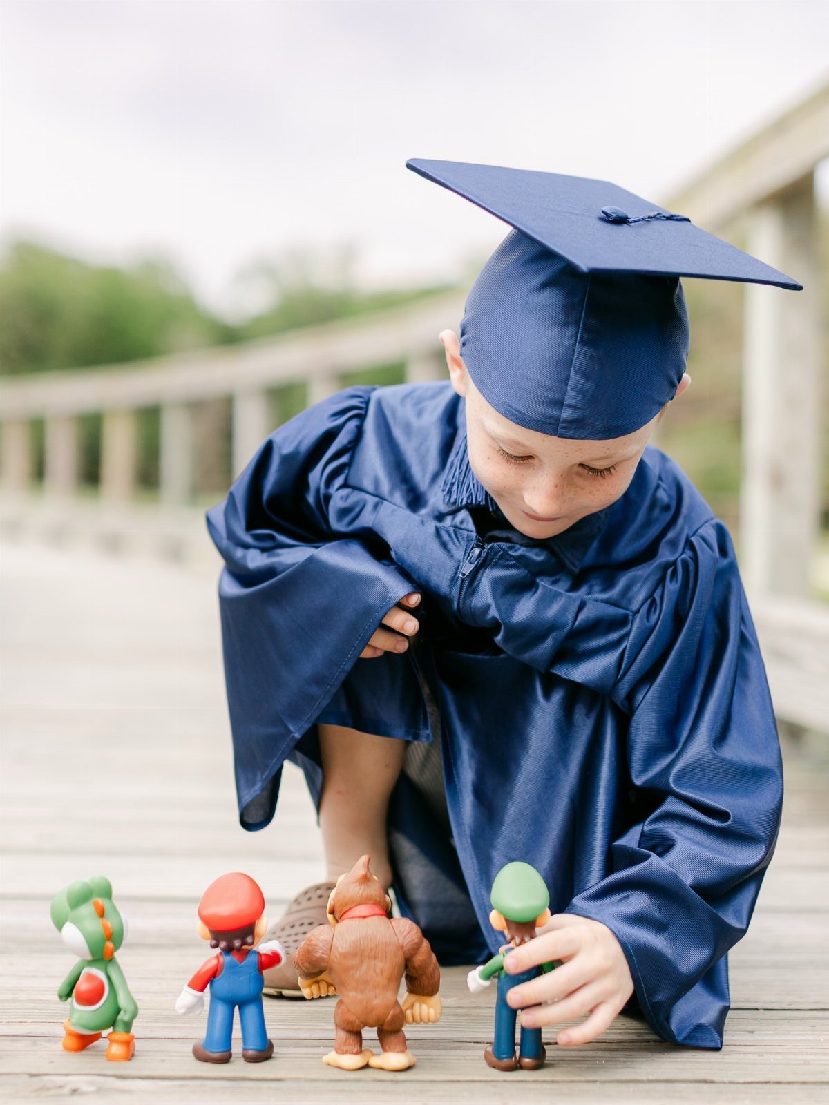 Boy in navy graduation gown and cap playing with Mario figurines on boardwalk