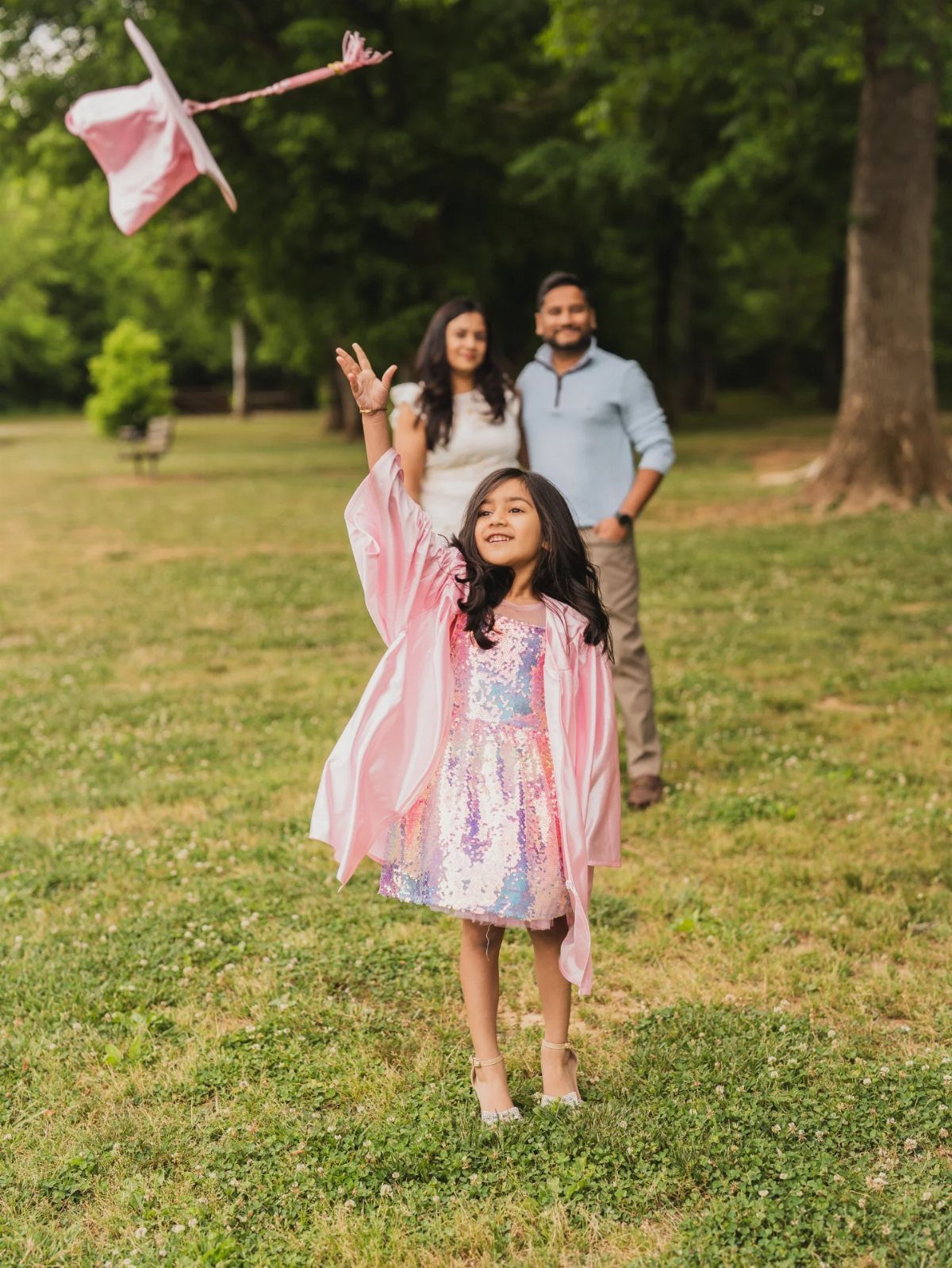 Young girl in pink graduation gown throwing her cap in a grassy park