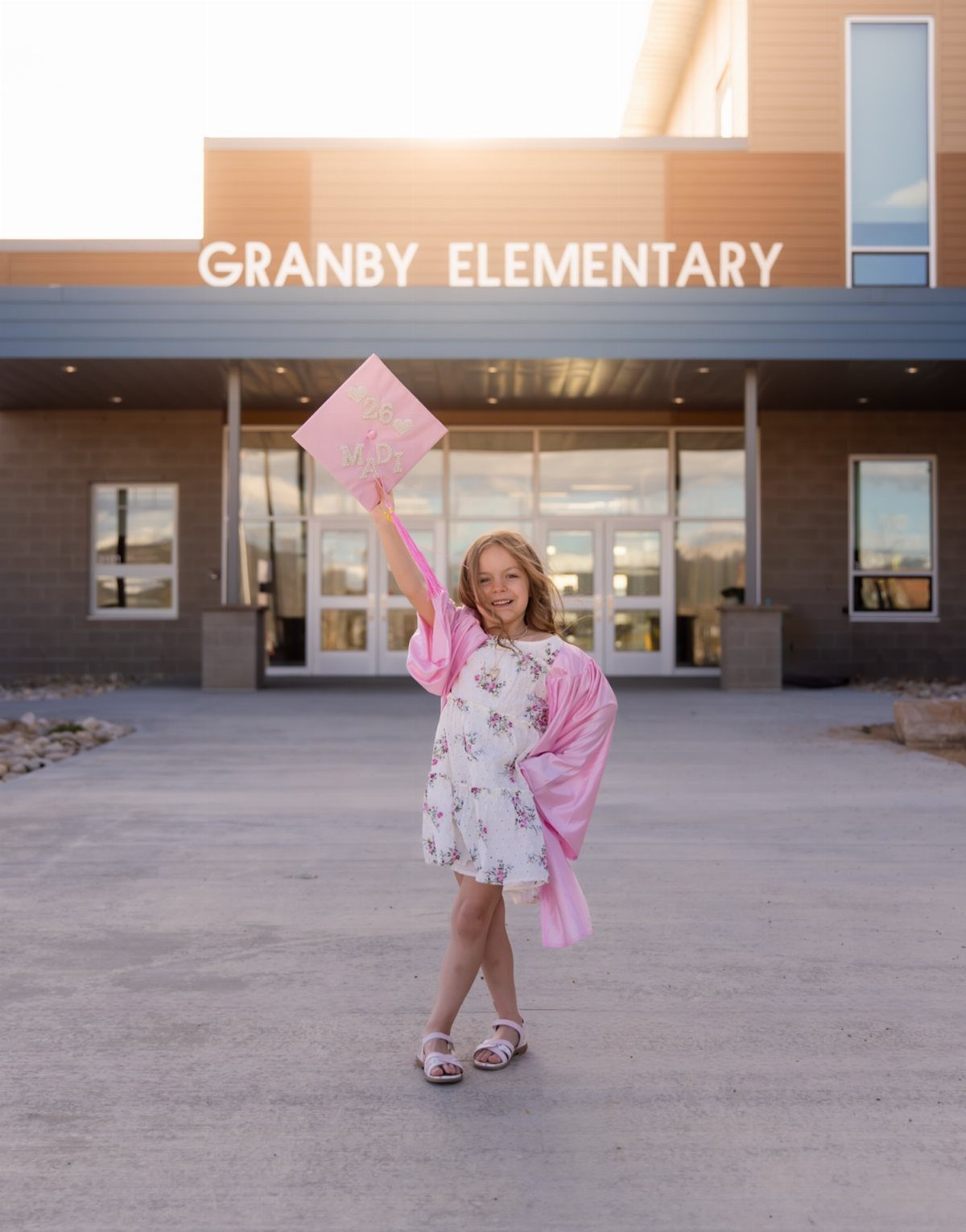 Young girl in pink graduation gown holding a cap before an elementary school