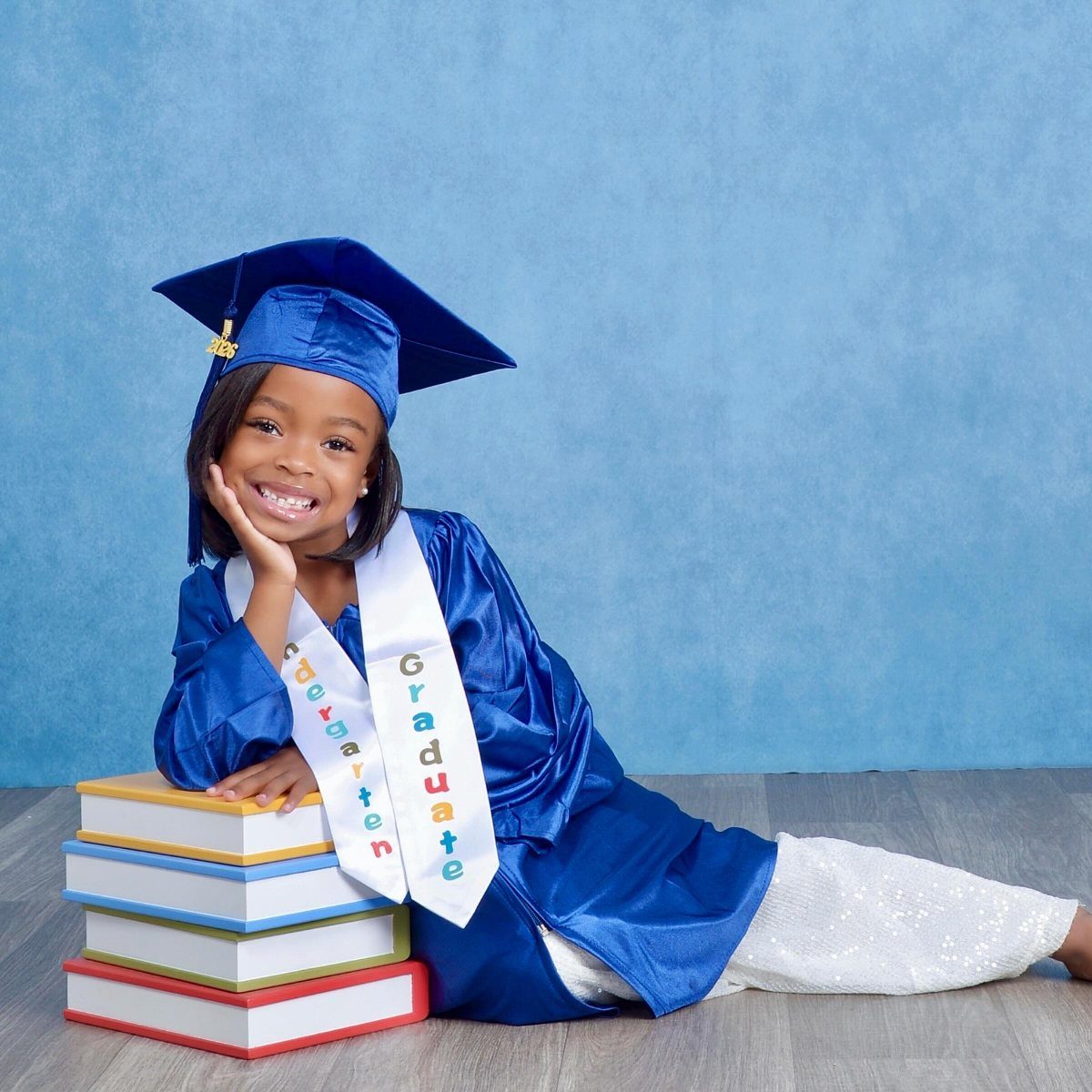 Young girl in blue graduation cap and gown leaning on colorful books
