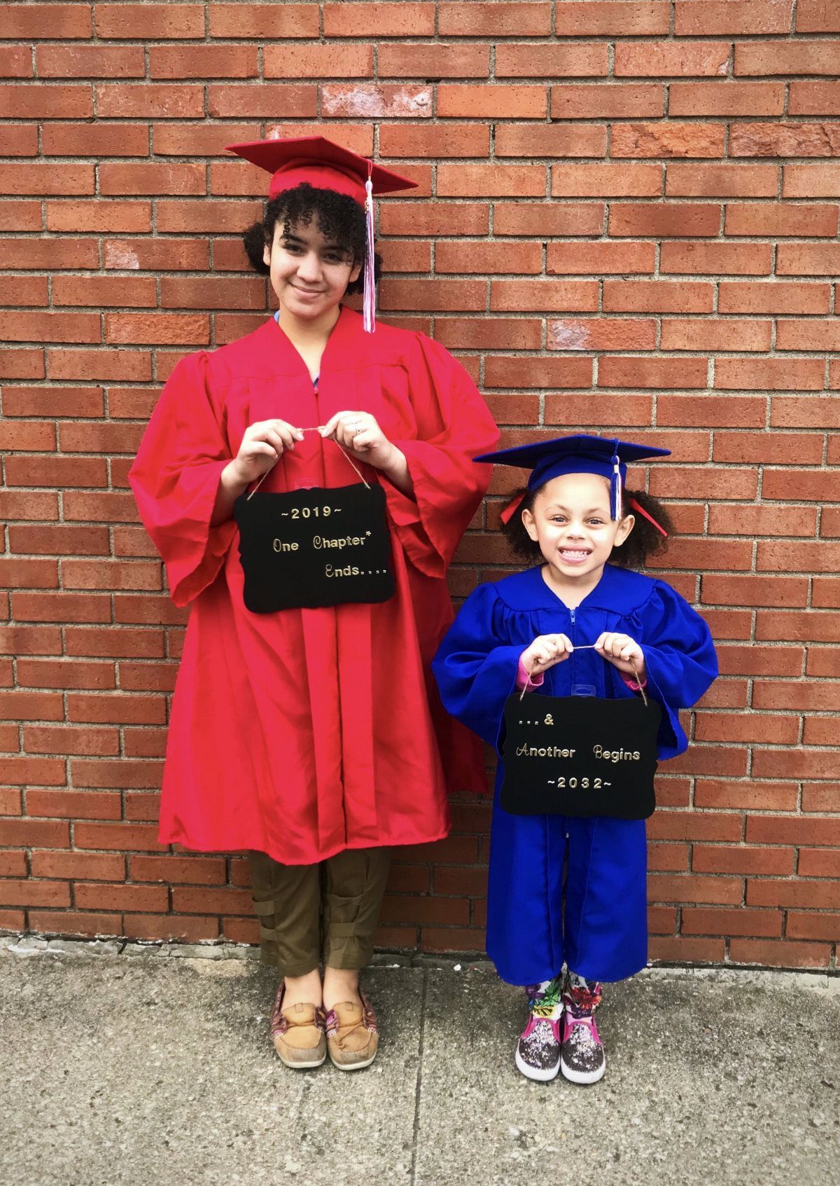 Two sisters in red and blue graduation gowns posing against a brick wall.