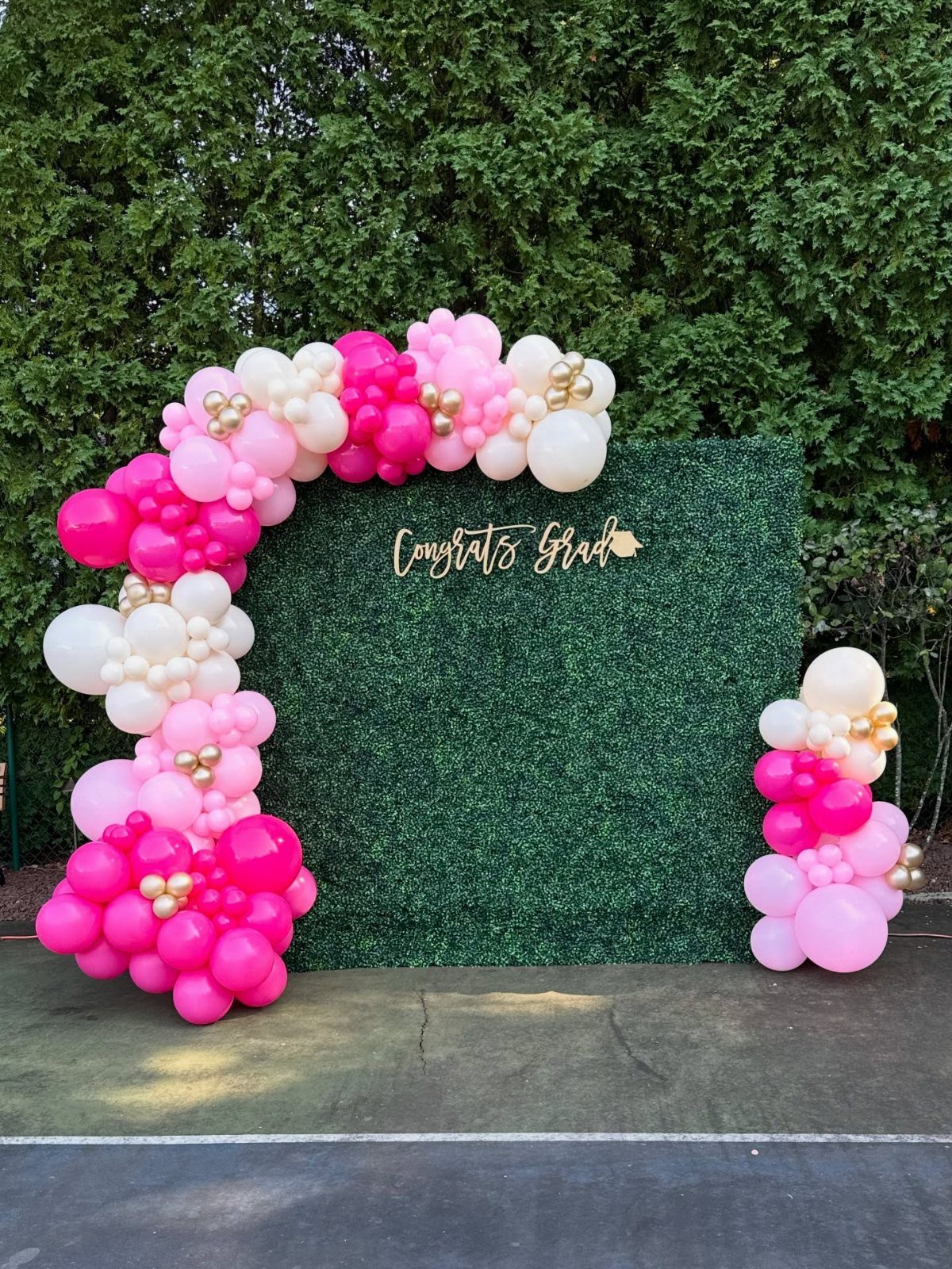 Green hedge backdrop wall with pink and white balloons and Congrats Grad sign.