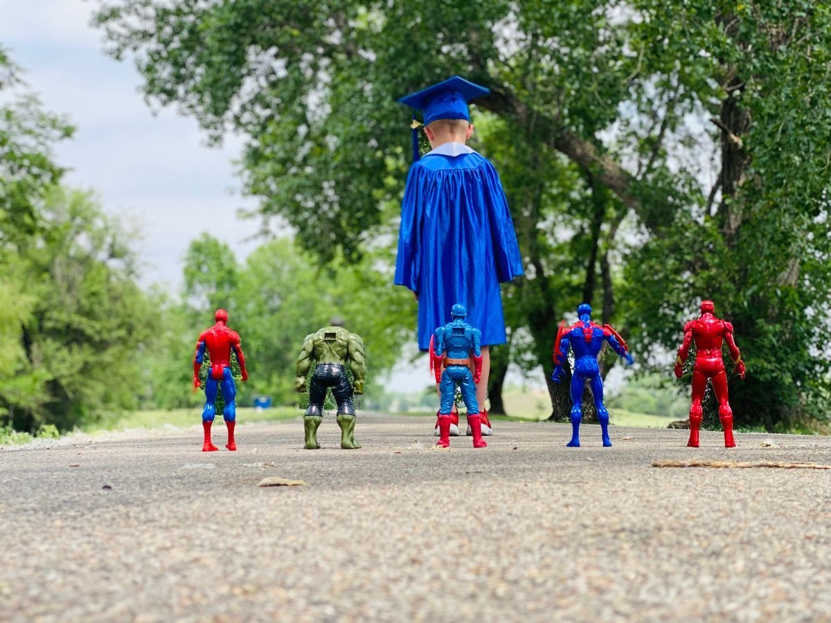 Child in blue graduation gown walking toward superhero action figures on a path