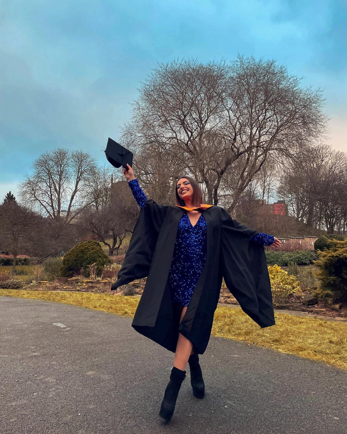 Woman in black graduation gown and blue dress holding up a graduation cap.